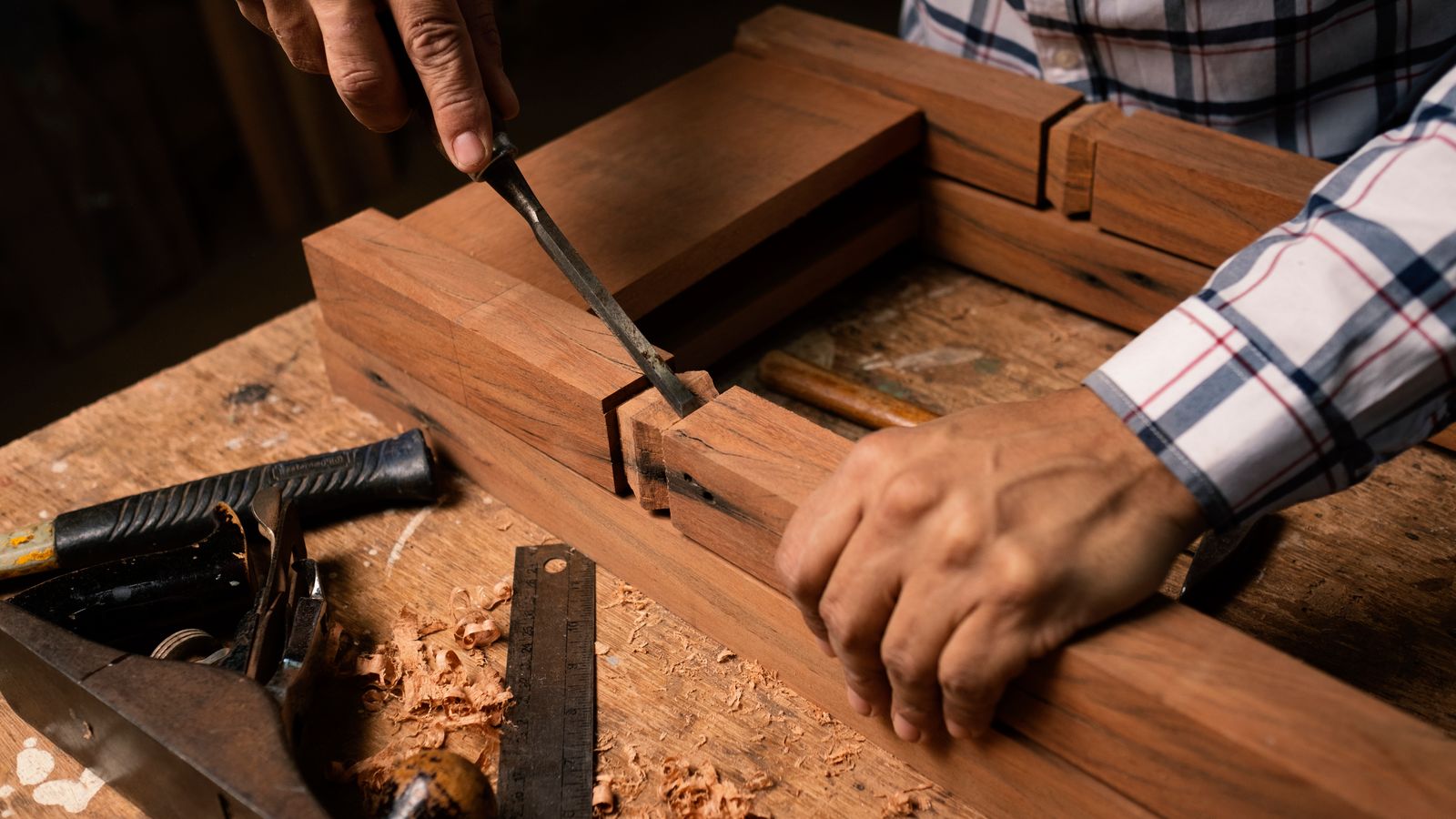 Close-up of a woodworker's hands guiding a chisel across a piece of timber, with a shallow depth of field.