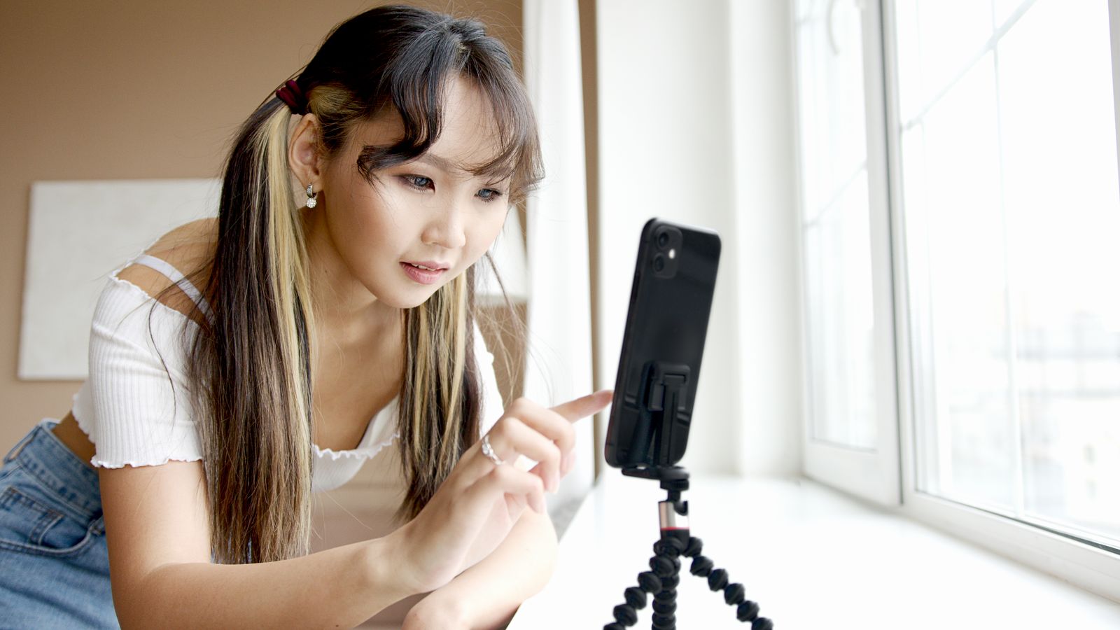 A woman in a white top recording with a smartphone mounted on a tripod beside a large window in a bright room.