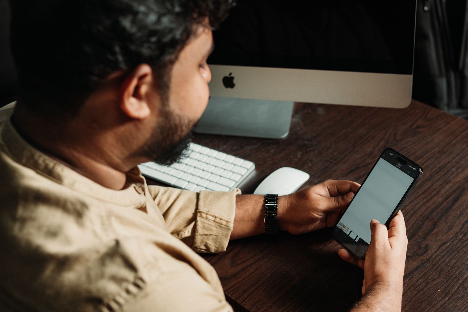 A man checking his smartphone at an office desk beside a computer monitor and keyboard.