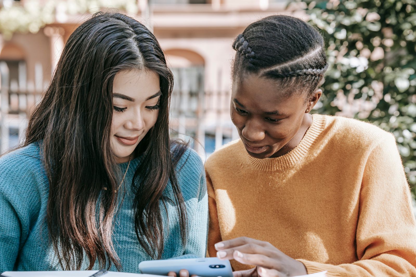 Three women standing outdoors on a street, smiling as they watch a video together on a smartphone.
