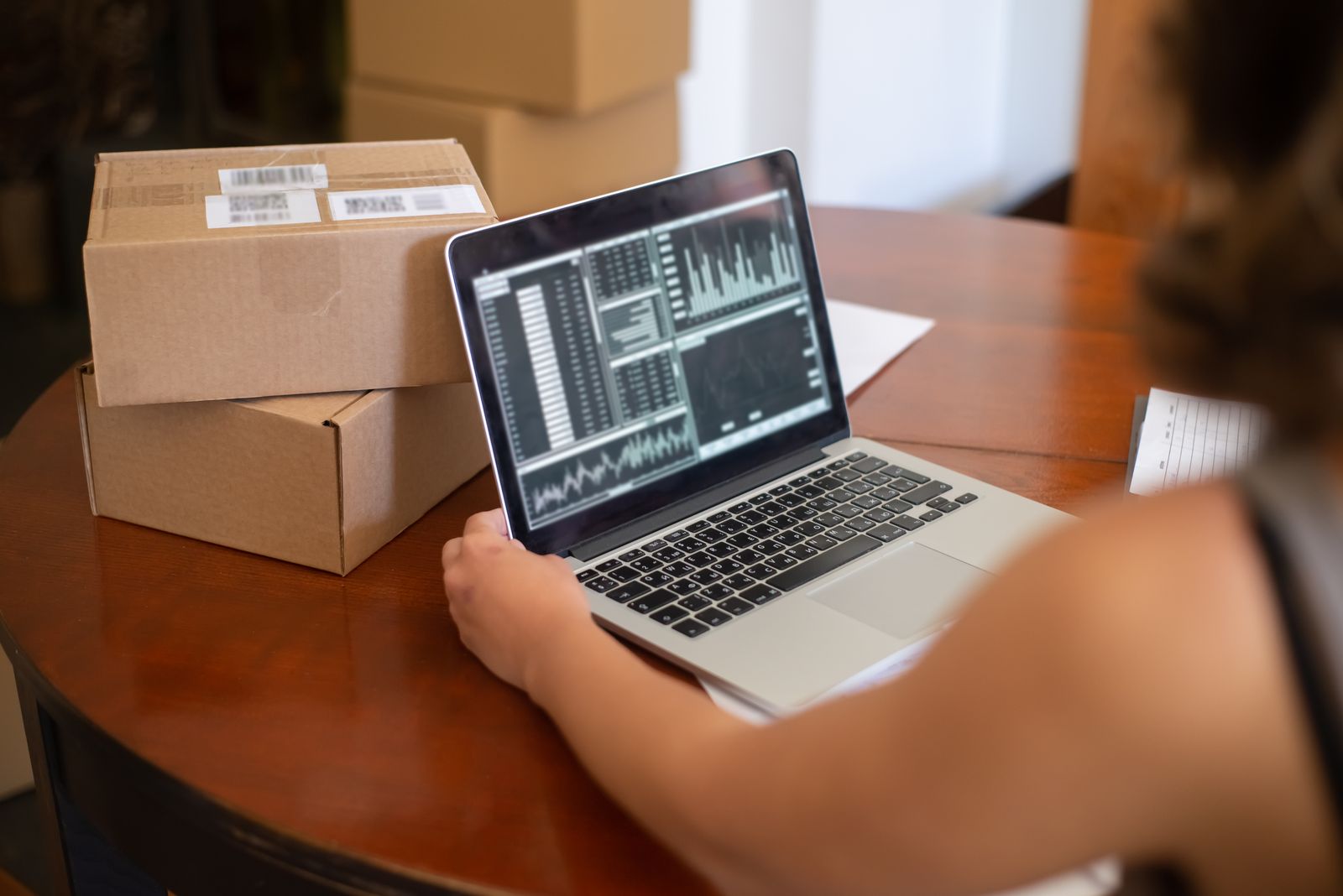 Small business owner sitting at a table surrounded by cardboard shipping boxes, studying charts and graphs on a laptop screen.