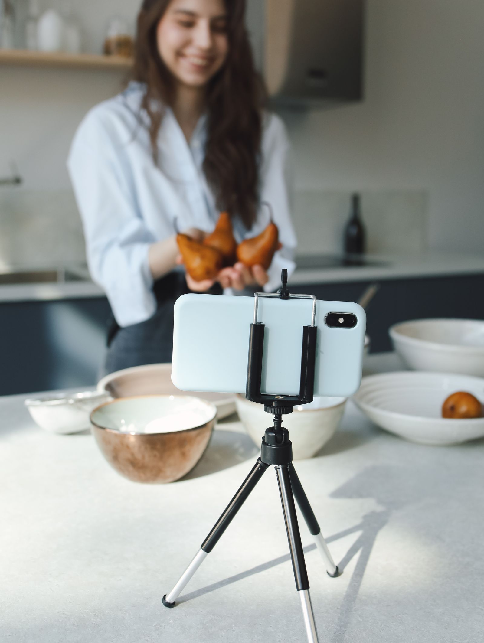A woman holding pears while recording a cooking video on a smartphone mounted on a tripod in a bright kitchen.
